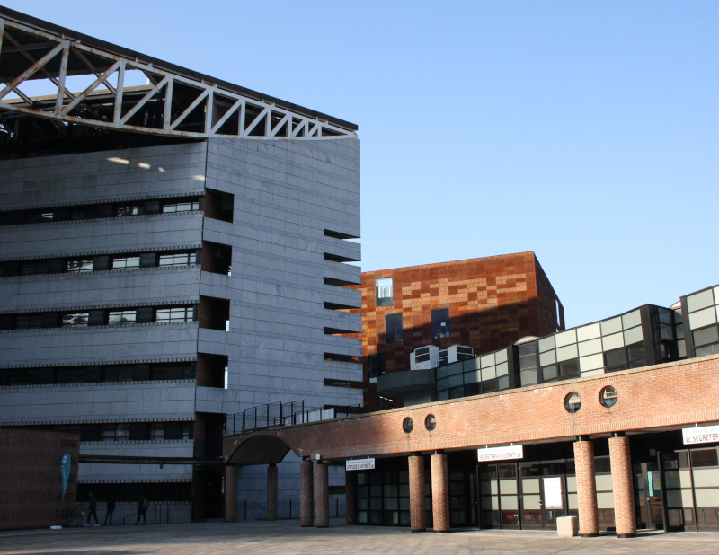 Edificio grigio, scorcio altro edificio color ruggine, porticato mattoni rossi, scorcio piazza